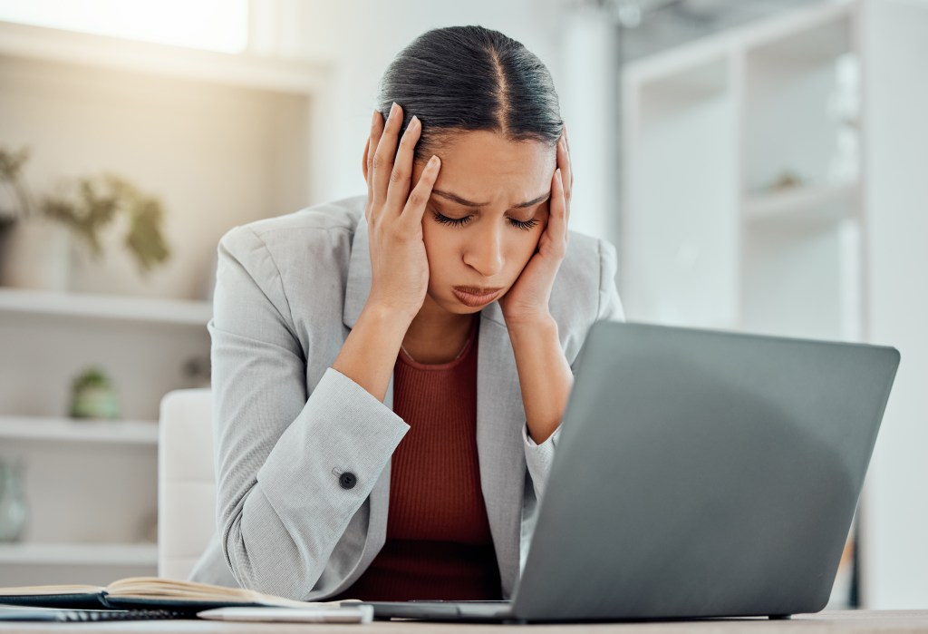 Stressed young businesswoman holding head in hand, worriedly looking at laptop